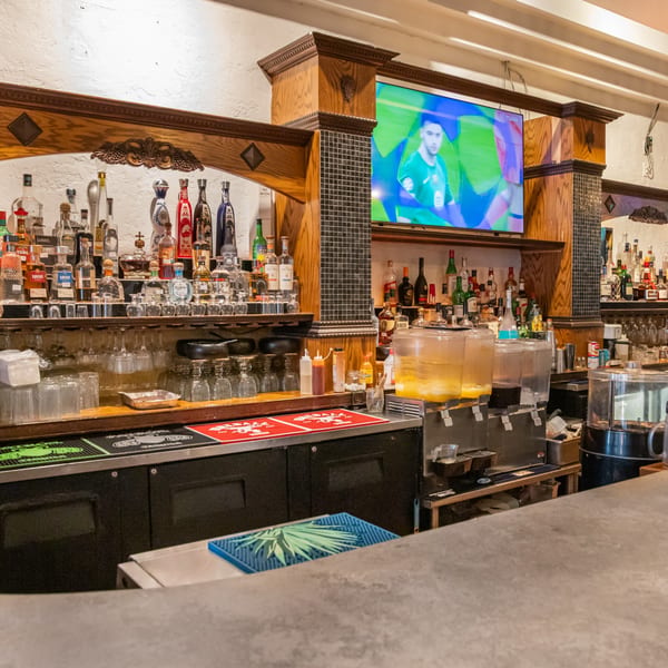 A well-stocked bar at San Jose Mexican Restaurant with various liquor bottles and a TV showing soccer.