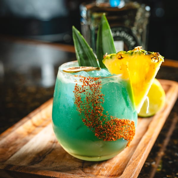 A vibrant blue cocktail with chili powder, pineapple, and lime on a wooden tray.