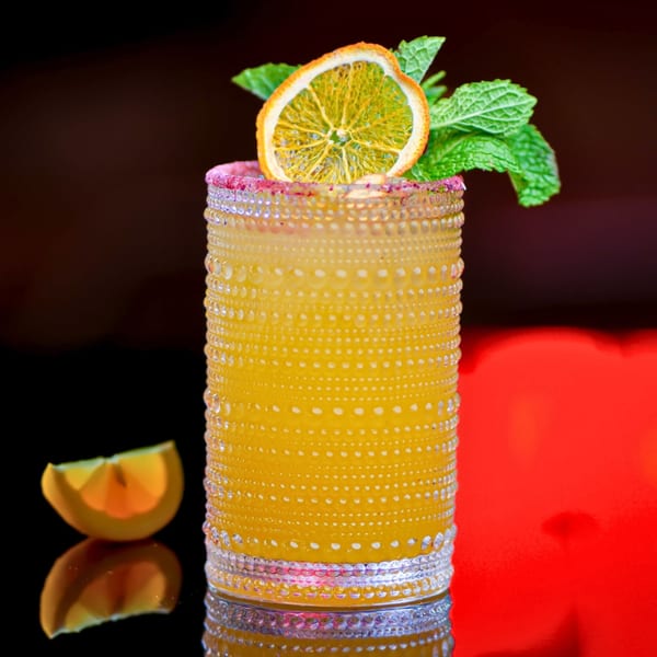 A vibrant yellow cocktail in a textured glass, garnished with a dried orange slice and mint leaves.