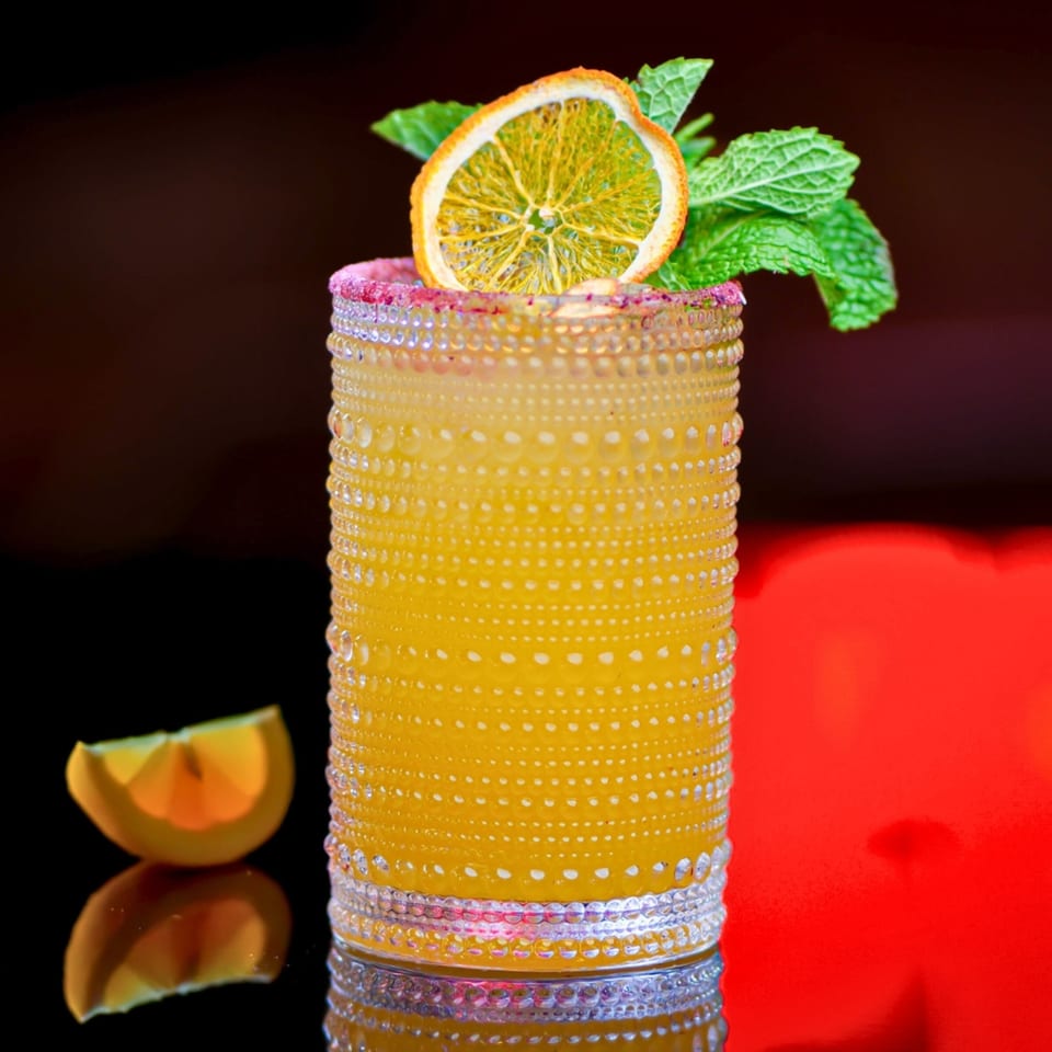 A vibrant yellow cocktail in a textured glass, garnished with a dried orange slice and mint leaves.