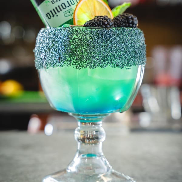 A blue and green margarita with a black sugar rim, garnished with blackberries, orange, and a beer bottle.