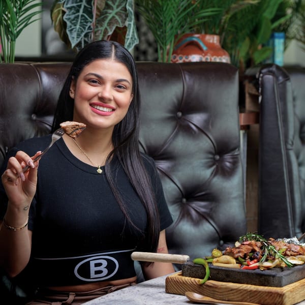 Smiling woman holding a piece of steak on a fork, with a sizzling fajita platter on the table.