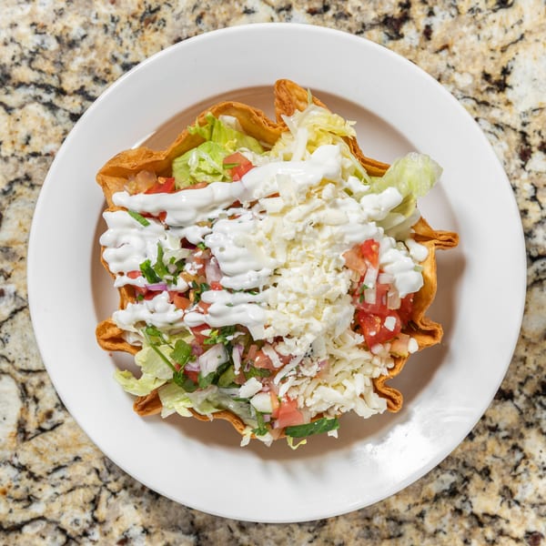 A taco salad in a crispy tortilla bowl, topped with lettuce, pico de gallo, sour cream, and cheese.