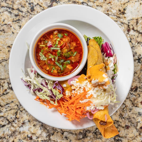 A white plate holds a bowl of red soup with cilantro, and taquitos with shredded cabbage and carrots.