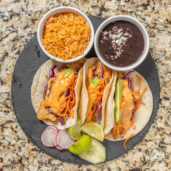 Three fish tacos with rice, black beans, radishes, limes, and cucumber on a dark plate.