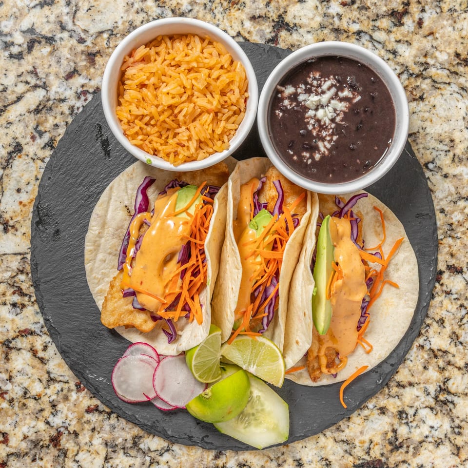 Three fish tacos with rice, black beans, radishes, limes, and cucumber on a dark plate.