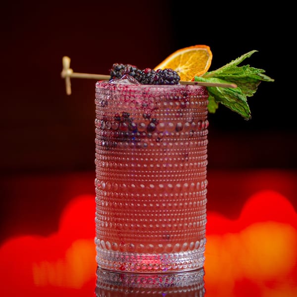 A vibrant red cocktail in a textured glass, garnished with blackberries, orange slice, and mint.
