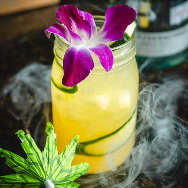 A vibrant yellow cocktail in a mason jar with cucumber, a purple orchid, and smoky dry ice.